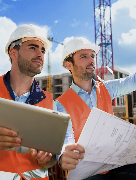 Zwei Bauingenieure mit Schutzhelmen betrachten Baupläne auf einer Baustelle.
