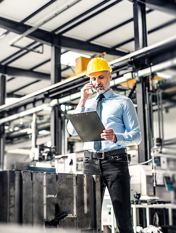 Ein Ingenieur mit Helm telefoniert in einer Industrieanlage und betrachtet dabei ein Dokument.