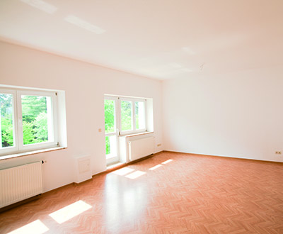 Ein leerer, heller Raum mit Parkettboden und Fenstern, durch die das Sonnenlicht scheint.
