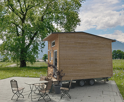 Ein kleines, mobiles Holzhaus steht auf einem Anhänger neben einer Sitzgruppe im Freien mit einem idyllischen, bergigen Hintergrund.